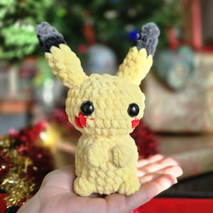 Puede incluir: Un juguete de peluche Pikachu amarillo tejido a crochet con orejas negras y mejillas rojas. El juguete tiene grandes ojos negros y está de pie sobre una mano.