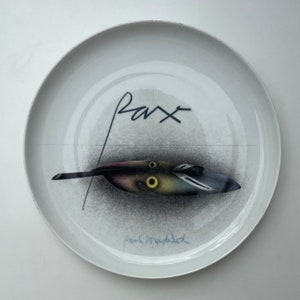 May include: A white ceramic plate with a black and white drawing of a feather with a yellow eye. The word "pax" is written in black above the feather. The plate is signed "Paul Klee" in black.