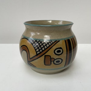 May include: A beige ceramic vase with a geometric design in brown, black, and blue. The vase has a rounded body and a wide opening.