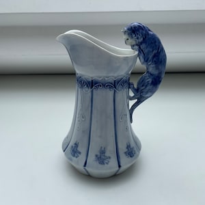 Puede incluir: Una jarra de porcelana azul y blanca con un asa de gato. El gato está encaramado en el borde de la jarra y mirando hacia abajo. La jarra tiene un diseño floral azul y blanco.