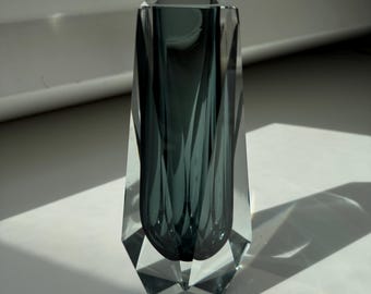 Könnte beinhalten: Eine hohe, facettierte Glasvase mit rechteckiger Öffnung. Die Vase hat eine rauchige teal Farbe und ein geometrisches Design. Das klare Glasäußere lässt Licht durch, wodurch Schatten entstehen. Die Vase wird wahrscheinlich zu dekorativen Zwecken verwendet.