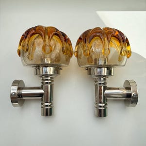 May include: Pair of vintage wall sconces. Each sconce features a clear glass globe with amber and brown accents, mounted on a polished chrome base. The sconces have a unique, decorative design, perfect for adding a touch of retro style to any room.