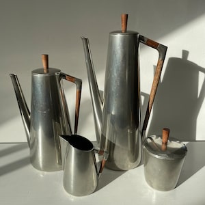 Puede incluir: Un juego de cuatro teteras de plata con asas de madera. Las teteras son de diferentes tamaños y formas, y todas están pulidas hasta brillar.