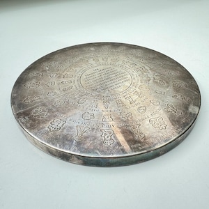 May include: A silver circular disc with a raised rim and intricate engravings. The engravings include a variety of symbols, numbers, and text in French. The text includes the words "Argent Premier" and "Pieces Terminees".