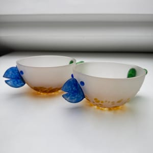 May include: Two frosted glass bowls with blue fish-shaped handles and yellow and orange speckled bottoms. The bowls have small blue dots and green accents. The bowls are on a white surface.