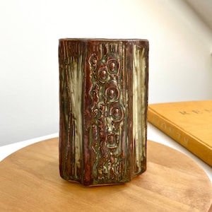 Peut inclure: Un vase en céramique brun et vert avec un motif texturé et en relief. Le vase a une forme hexagonale et une ouverture légèrement évasée.
