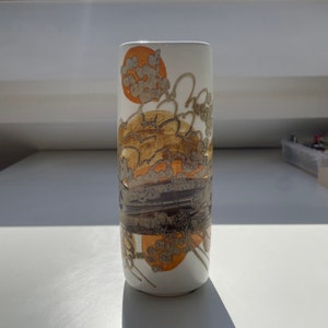 Può includere: Vaso in ceramica bianca con un design astratto marrone e arancione. Il vaso ha una forma cilindrica e una finitura liscia e lucida.