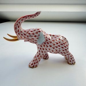 May include: A ceramic elephant figurine with a red and white pattern. The elephant has gold-colored tusks and feet, with light blue accents on the ears. The trunk is raised, and the figurine is on a white surface.