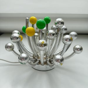 May include: A vintage chrome and glass Sputnik-style table lamp. The lamp features a circular base with multiple curved arms extending outwards, each holding a silver orb. Some arms have yellow or green spheres. A power cord extends from the base.