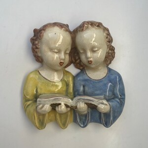 May include: A ceramic wall hanging depicting two figures with closed eyes, reading a book. One figure wears a yellow garment, the other a blue garment. The figures have brown curly hair and are reading a book with a white cover.