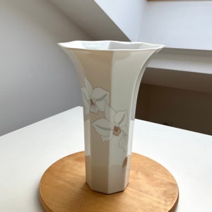 May include: White ceramic vase with a geometric shape and a floral design featuring white orchids with brown and green accents.