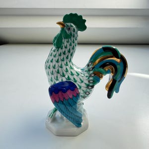 May include: A ceramic rooster figurine with a green comb and tail feathers in shades of blue, pink, and gold. The body is white with a green scale pattern. The rooster stands on a white hexagonal base.