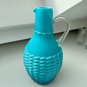 May include: A turquoise blue glass jug with a clear glass handle. The jug has a rope-like design around the body.