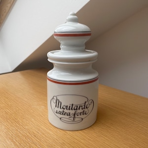 Könnte beinhalten: Weiße Keramik-Senfdose mit einem roten und schwarzen Streifen am oberen Rand und den Worten "Moutarde extra forte" in schwarzer Schrift.