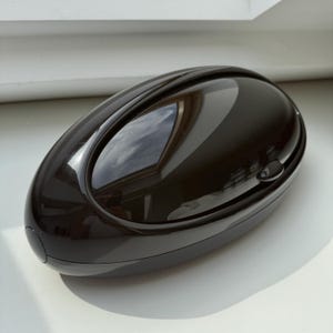 Może przedstawiać: Czarne, owalne, twarde plastikowe etui o gładkim, błyszczącym wykończeniu. Etui ma mały, okrągły przycisk z boku.