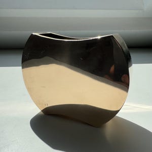 Peut inclure: Un vase en métal doré poli, de forme incurvée unique. Le vase a une large ouverture et une base arrondie. La surface réfléchit la lumière, créant des reflets et des ombres. C'est un objet décoratif.