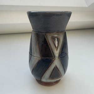 Peut inclure: Un petit vase en céramique avec un motif géométrique dans les tons de bleu, gris et marron. Le vase a un corps arrondi et une ouverture évasée.