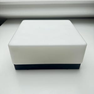 May include: A square, two-tone box with a white top and a black base. The box has a clean, minimalist design, with a smooth, matte finish. The box is sitting on a white surface.