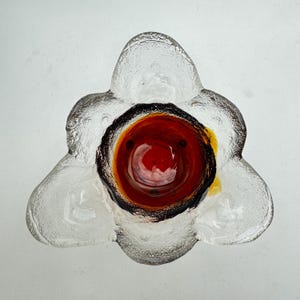 Pode incluir: Uma tigela de vidro transparente em forma de flor com um design central vermelho e preto. A tigela tem três pétalas arredondadas e uma superfície texturizada. O centro apresenta um tom vermelho vibrante com detalhes em preto e amarelo.