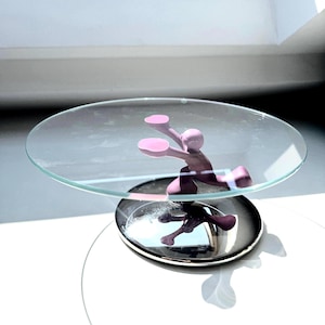 Op de afbeelding: Een decoratieve taartstandaard met een heldere glazen bovenkant en een chromen voet. Een roze, gestileerde figuur is op de standaard geplaatst, wat een grillige toets toevoegt. De standaard is ontworpen om desserts of andere items te presenteren.