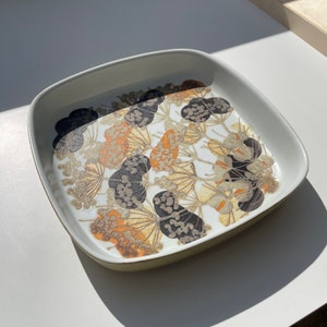 Può includere: Un piatto da portata quadrato in ceramica con sfondo bianco e un motivo floreale in tonalità di marrone, nero e arancione.