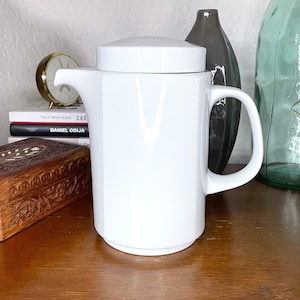 May include: A white ceramic teapot with a lid and a handle. The teapot is sitting on a wooden surface.