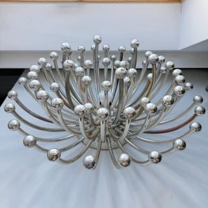 Puede incluir: Una lámpara de araña Sputnik plateada, de diseño moderno de mediados de siglo. Cuenta con múltiples brazos curvos que irradian desde un punto central, cada uno terminando en una bombilla esférica. La lámpara está montada sobre una superficie blanca.