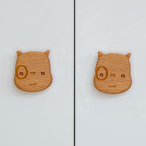 May include: Two wooden cabinet knobs shaped like animal faces with a simple design. The knobs are light brown and have a slightly textured surface.
