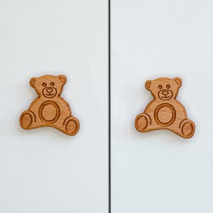 Peut inclure: Deux boutons de porte en bois en forme d'ours avec un design gravé simple. Les boutons sont de couleur brun clair et sont fixés à une porte de placard blanche.