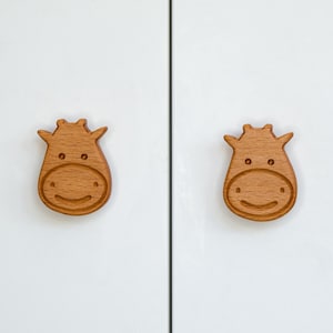 May include: Two wooden cow-shaped cabinet knobs with a smiling face. The knobs are light brown and have a smooth finish.