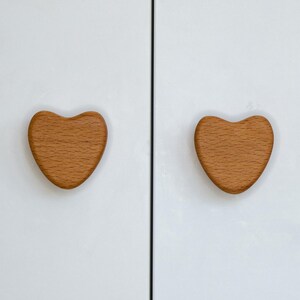 Peut inclure: Deux boutons de placard en bois en forme de cœur. Les boutons sont de couleur brun clair et sont montés sur une porte d'armoire blanche. Les boutons sont en bois et ont une finition lisse. La porte de l'armoire est blanche et a un aspect épuré et moderne.