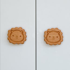 Puede incluir: Dos pomos de armario de madera con un diseño de cara de oso tallado. Los pomos son de color marrón claro y tienen una forma ligeramente redondeada.