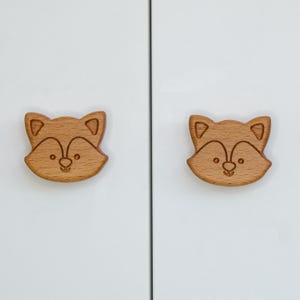 Puede incluir: Dos pomos de armario de madera con forma de cara de zorro. Los pomos son de color marrón claro y tienen un diseño simple y tallado.