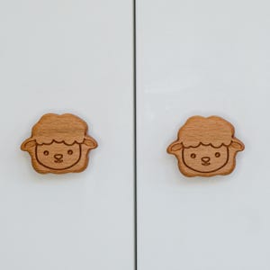 Op de afbeelding: Twee houten kastknoppen in de vorm van schapenkoppen. De knoppen zijn lichtbruin en hebben een eenvoudig, cartoonachtig ontwerp.