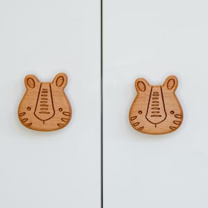 Może przedstawiać: Dwa drewniane gałki do szafek w kształcie twarzy tygrysa. Gałki są jasnobrązowe i mają szczegółowy wzór twarzy tygrysa.