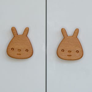 May include: Two wooden bunny-shaped cabinet knobs with a light brown finish. The knobs have a simple, cartoon-like face with two small eyes and a nose.