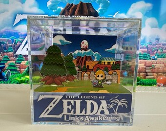 May include: A clear acrylic cube display case with a pixelated 8-bit scene from The Legend of Zelda: Link's Awakening video game. The scene features Link, a volcano, and a town. The case has a blue base with the game's title and logo.