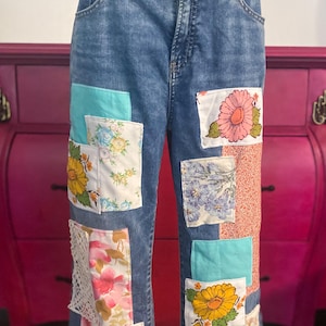 May include: Blue denim jeans with colorful floral patches. Patches include various floral patterns in pink, yellow, and blue, with a touch of lace. The jeans have a classic five-pocket design.