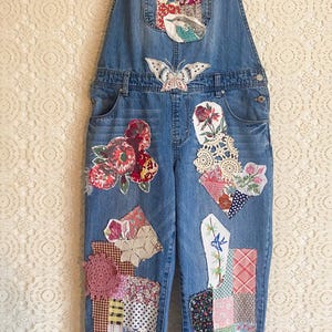 May include: Light blue denim overalls with decorative patches. Patches include floral, butterfly, and geometric designs in various colors. The overalls have a bib pocket and adjustable straps.