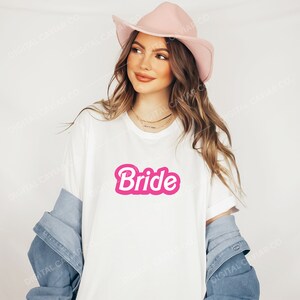 May include: A white t-shirt with a pink "Bride" graphic in a retro font. The shirt is being worn by a person wearing a pink hat and a denim jacket.