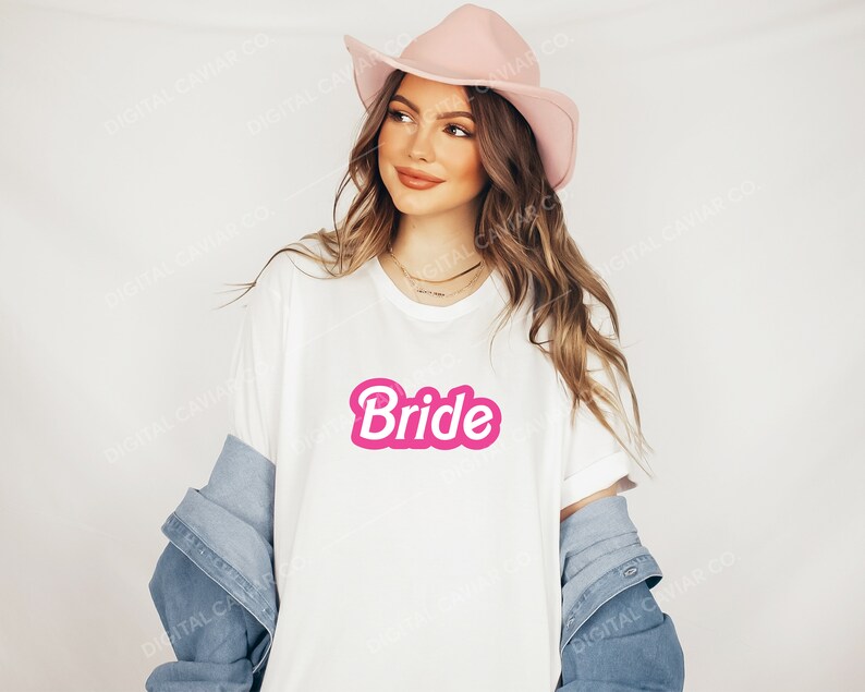 May include: A white t-shirt with a pink "Bride" graphic in a retro font. The shirt is being worn by a person wearing a pink hat and a denim jacket.