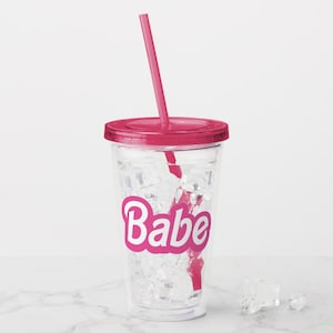 May include: A clear plastic tumbler with a pink lid and straw. The tumbler has ice cubes and the word "Babe" printed on the side in pink letters.