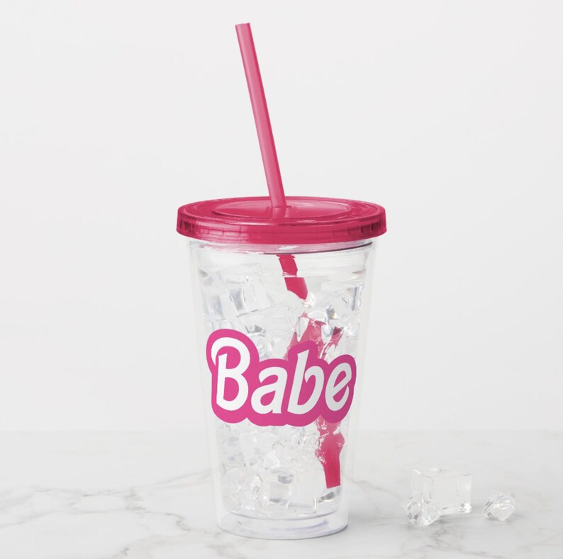 May include: A clear plastic tumbler with a pink lid and straw. The tumbler has ice cubes and the word "Babe" printed on the side in pink letters.