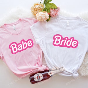 May include: Two pink and white t-shirts with the words "Babe" and "Bride" in pink letters. The shirts are laid out on a white fuzzy surface with a bottle of champagne.
