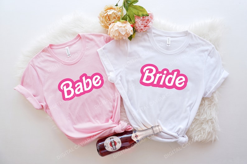 May include: Two pink and white t-shirts with the words "Babe" and "Bride" in pink letters. The shirts are laid out on a white fuzzy surface with a bottle of champagne.