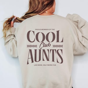 Könnte beinhalten: Ein cremefarbenes Sweatshirt mit dem Aufdruck "PROUD MEMBER OF THE COOL CLUB AUNTS LIKE MOM, ONLY MORE FUN" in braunen Buchstaben. Das Sweatshirt hat einen Rundhalsausschnitt und lange Ärmel. In der unteren rechten Ecke des Bildes steht "SWEATER WHITE".