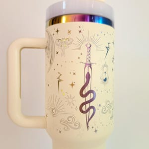 May include: A white travel mug with a gold iridescent lid and a gold design of a snake wrapped around a sword, stars, and other mystical symbols.