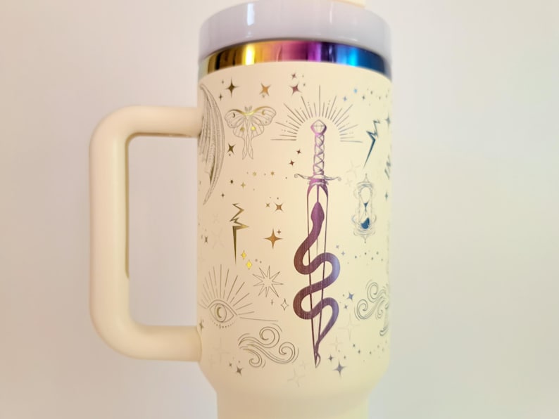 May include: A white travel mug with a gold iridescent lid and a gold design of a snake wrapped around a sword, stars, and other mystical symbols.