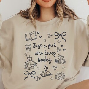 Könnte beinhalten: Sandfarbener Pullover mit dem Schriftzug "Just a girl who loves books" in schwarzer Schrift. Das Design umfasst Illustrationen von Büchern, Kaffeetassen, Schleifen und Herzen. Der Pullover hat eine neutrale Farbe.