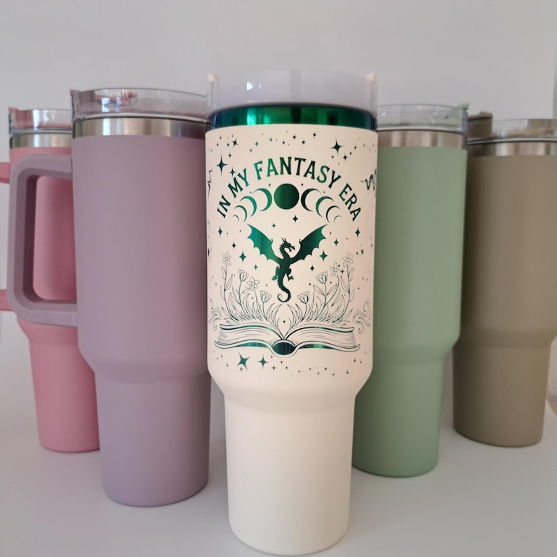 May include: Four stainless steel tumblers with different colored lids and bodies. The tumbler in the center has a white body with a green dragon and the text "In My Fantasy Era" printed on it.
